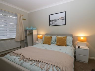 A bedroom with a bed with two mustard cushions bedside tables with lamps an open wardrobe with folded towels and hangers and a window with blinds and curtains at Sandown in Bowness-on-Windermere