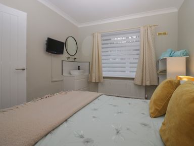 A bedroom with a bed with pillows a window with curtains a television on the wall a round mirror a sink a cabinet and shelves with towels at Sandown in Bowness-on-Windermere