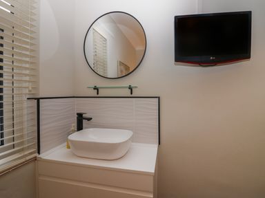 A bathroom with a white sink and black faucet a round mirror and a wall-mounted TV at Sandown in Bowness-on-Windermere