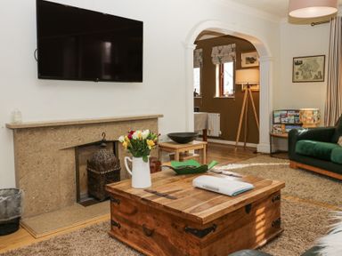 A living room with a wooden coffee table flowers on it a mounted TV and an arched doorway at Sandown in Bowness-on-Windermere