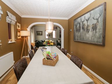 A dining room with a long table covered with a cloth and six chairs a basket with snacks and a wine bottle and flowers on the table a large painting of deer on the wall at Sandown in Bowness-on-Windermere