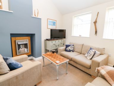 A living room with two beige sofas a wooden coffee table a fireplace a television on a cabinet and windows at Driftwood Apartment in Amble-by-the-Sea