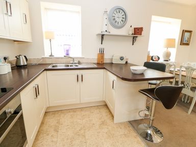 A kitchen with white cabinets a double sink a dark countertop a bar stool and a dining area with chairs at Driftwood Apartment in Amble-by-the-Sea