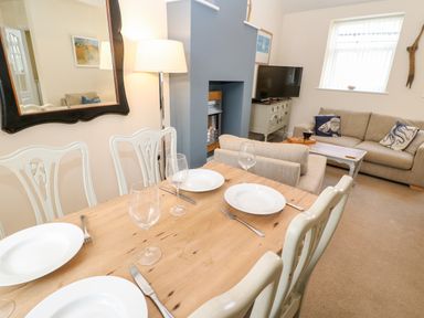 A dining table set with plates and glasses next to a living room with sofas a coffee table a television and a fireplace at Driftwood Apartment in Amble-by-the-Sea