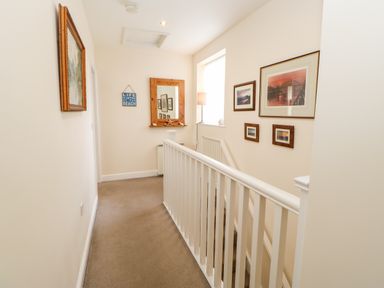 A hallway with beige carpet a wooden framed mirror pictures on walls and white stair railing at Driftwood Apartment Amble-by-the-Sea