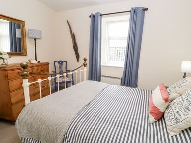 A bedroom with a bed with striped bedding a wooden dresser with a lamp and mirror a blue chair and a window with blue curtains at Driftwood Apartment in Amble-by-the-Sea