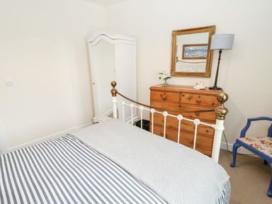 A bedroom with a bed striped bedspread a wooden chest of drawers a white wardrobe a mirror floor lamp and a blue chair at Driftwood Apartment in Amble-by-the-Sea