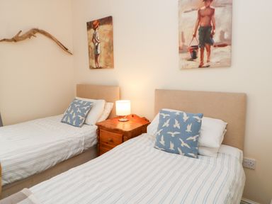 A bedroom with two single beds with striped bedding and bird-patterned pillows a wooden nightstand with a lamp and paintings above the beds at Driftwood Apartment in Amble-by-the-Sea
