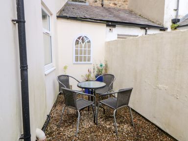 An outdoor seating area with a glass table and four chairs on a gravel floor at Driftwood Apartment in Amble-by-the-Sea