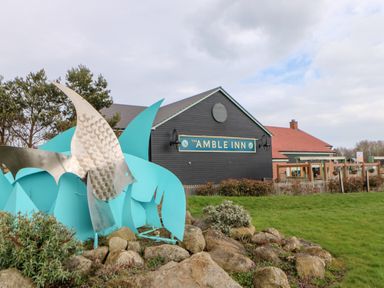 Metal fish sculpture with blue fins on rocks and grass outside a building with a sign that reads The Amble Inn