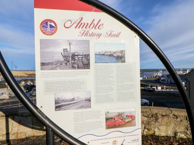 An information board about Amble History Trail near a harbor with water and boats in the background