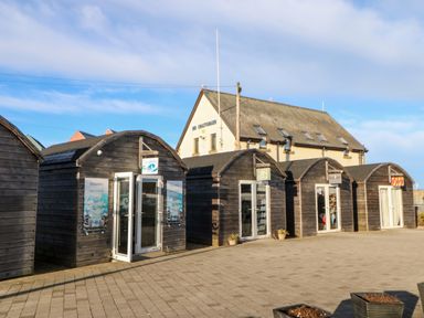 A row of small wooden shops with signs and glass doors in a paved area near a large building at Driftwood Apartment in Amble-by-the-Sea