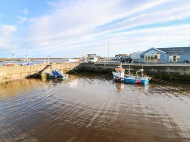 A harbor with two small boats moored alongside a concrete wall and buildings in the background at Driftwood Apartment in Amble-by-the-Sea