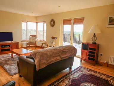 A living room with a sofa covered by a blanket facing a wooden coffee table and two armchairs near windows a television on a wooden stand a wooden cabinet with a lamp and a door leading to a balcony at Bluebell House and Gardens Gurteenulla near Ballydehob County Cork