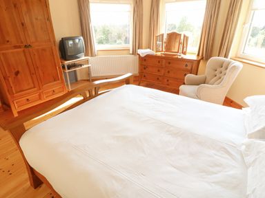 A bedroom with a wooden bed a wooden wardrobe a TV on a small table a wooden dresser with a mirror and a beige upholstered armchair at Bluebell House and Gardens Gurteenulla near Ballydehob County Cork