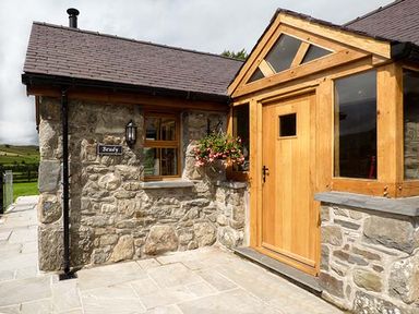 The exterior of a stone house with a wooden door and frame and flowers hanging by a window at Beudy in Llangwm near Bala