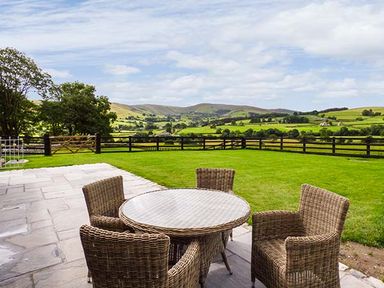 An outdoor patio with wicker chairs and a round glass table overlooking green fields and hills at Beudy in Llangwm near Bala