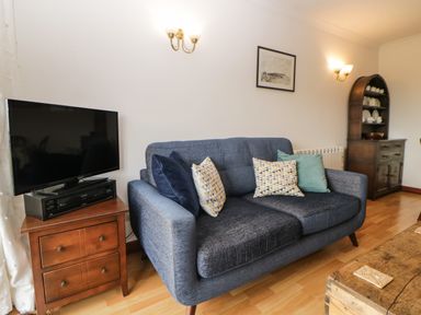 A living room with a blue sofa with cushions a wooden TV stand with a television and a wooden cabinet with shelves at Broom Cottage in Stoneleigh Holiday Village near Sidmouth
