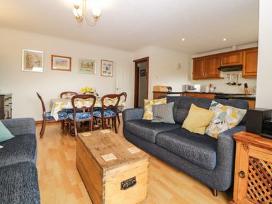 A living room with two blue sofas a wooden chest coffee table a dining table with six chairs and wooden kitchen cabinets at Broom Cottage Stoneleigh Holiday Village near Sidmouth