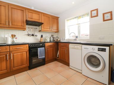 A kitchen with wooden cabinets a black stove a dishwasher a washing machine and a window at Broom Cottage Stoneleigh Holiday Village near Sidmouth