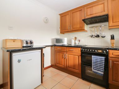 A kitchen with wooden cabinets a black stove a microwave a toaster a small white fridge and utensils hanging on the wall at Broom Cottage in Stoneleigh Holiday Village near Sidmouth