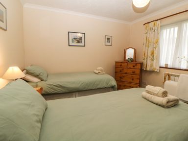 A bedroom with two beds a wooden chest of drawers and a window with floral curtains at Broom Cottage Stoneleigh Holiday Village near Sidmouth