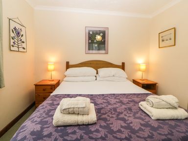 A bedroom with a double bed two bedside tables with lamps folded towels on the bed and framed pictures on the walls at Broom Cottage in Stoneleigh Holiday Village near Sidmouth