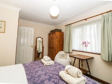 A bedroom with a bed covered with folded towels a wooden wardrobe a wooden table a chair with a cushion a full-length mirror and a window with curtains at Broom Cottage Stoneleigh Holiday Village near Sidmouth