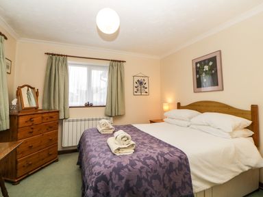 A bedroom with a double bed covered with a purple patterned blanket and towels on top next to a wooden chest of drawers with a mirror and a window with green curtains at Broom Cottage Stoneleigh Holiday Village near Sidmouth