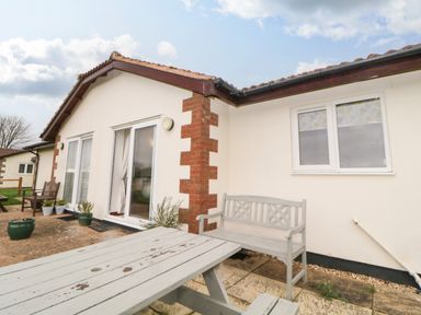 An outdoor patio area with a wooden picnic table and bench outside a single story house at Broom Cottage in Stoneleigh Holiday Village near Sidmouth