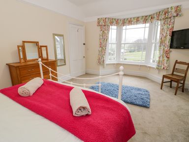 A bedroom with a bed covered by a red blanket and two rolled towels a wooden dresser with a mirror floral curtains on a bay window a wooden chair and a wall-mounted television at The Farm House in Bala