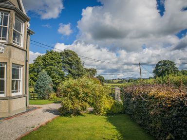 A garden with bushes and trees next to a building and a gravel path at The Farm House in Bala