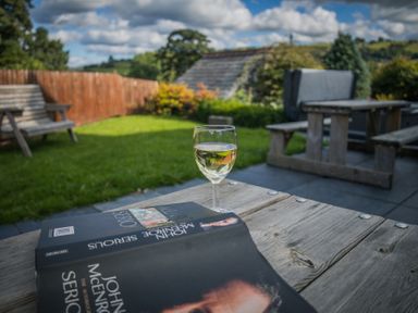 A glass of white wine and a book on a wooden table in a garden with benches at The Farm House in Bala