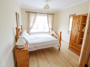 A bedroom with a wooden bed a nightstand with a lamp wooden wardrobe and a window with curtains at Shraigh Beach in Belmullet County Mayo