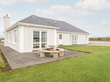 A single story house with white walls and dark roof a round stone outdoor table with curved benches on a concrete patio surrounded by grass at Shraigh Beach in Belmullet County Mayo
