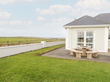A patio area with stone picnic table and curved benches next to a white house with glass doors and a garden with green grass at Shraigh Beach in Belmullet County Mayo
