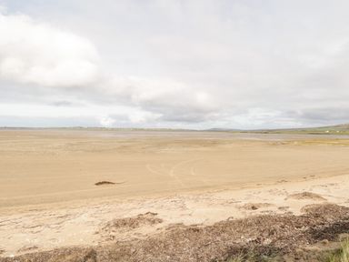A wide sandy beach with seaweed along the shore and a distant view of land under a cloudy sky at Shraigh Beach in Belmullet County Mayo