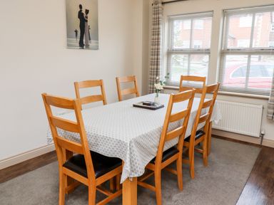A dining room with a wooden table covered by a patterned tablecloth surrounded by six wooden chairs near windows with blinds and a radiator at Darling Cottage Bamburgh