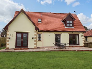 The back view of a house with a red tiled roof a patio with a table and chairs and a green lawn at Darling Cottage in Bamburgh