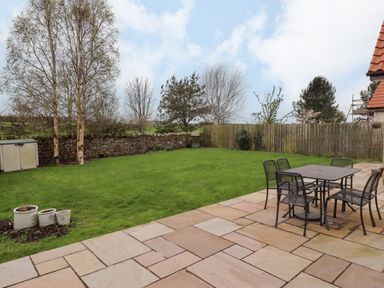 A garden with green grass a stone wall wooden fence trees patio with a table and four chairs at Darling Cottage in Bamburgh