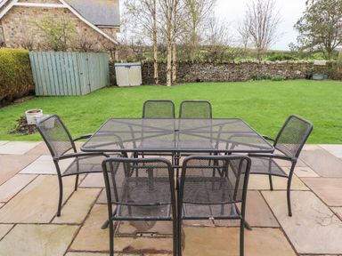 A patio with a metal table and six chairs overlooking a lawn with trees and a stone wall at Darling Cottage in Bamburgh