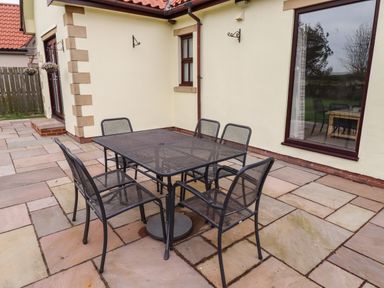A patio with a black metal table and six matching chairs outside a building with beige walls and red roof tiles at Darling Cottage in Bamburgh