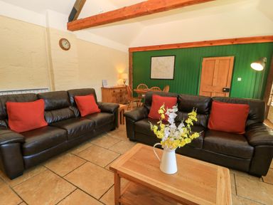 A living room with two leather sofas with red cushions a wooden coffee table with flowers and a dining table in the background at Keepers Cottage in Winterborne Stickland