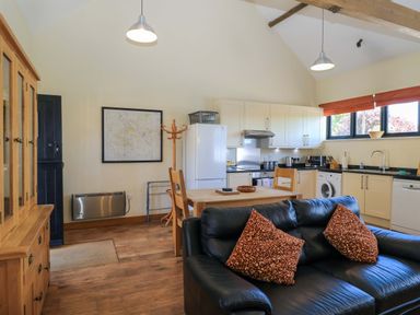 An open-plan kitchen and living area with a black sofa and patterned cushions a wooden dining table and chairs kitchen cabinets appliances and windows at The Old Cart Shed in Winterborne Stickland