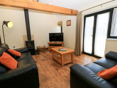 A living room with black sofas orange cushions a wood stove TV on a wooden stand and wooden coffee table at Rowdens Barn in Winterborne Stickland