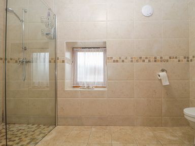 A tiled bathroom with a walk in shower a small window with curtains and a toilet paper holder at The Old Barn in Harman's Cross near Swanage