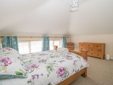A bedroom with a bed with floral bedding a wooden dresser a wicker chair and blue curtains at The Old Barn in Harman's Cross near Swanage