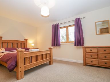 A bedroom with a wooden bed frame purple bedding a wooden dresser and a window with purple curtains at The Old Barn in Harman's Cross near Swanage