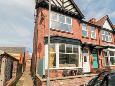 A red brick end terrace house with bay window yellow curtains and black metal chairs on the front patio at The Robins in Filey
