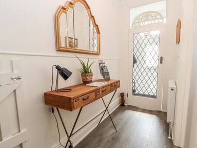 A hallway with a wooden console table holding a lamp plant and brochures below a gold-framed mirror and a glass door with a diamond pattern at The Robins in Filey